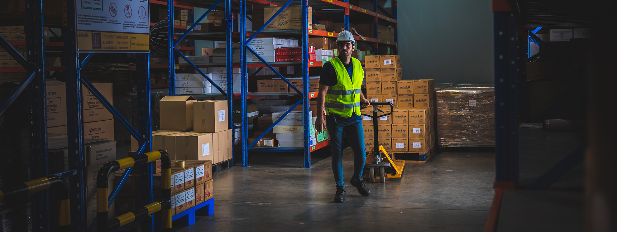 Warehouse worker in a high-visibility vest and white hard hat pulling a pallet truck between racks of boxed stock
