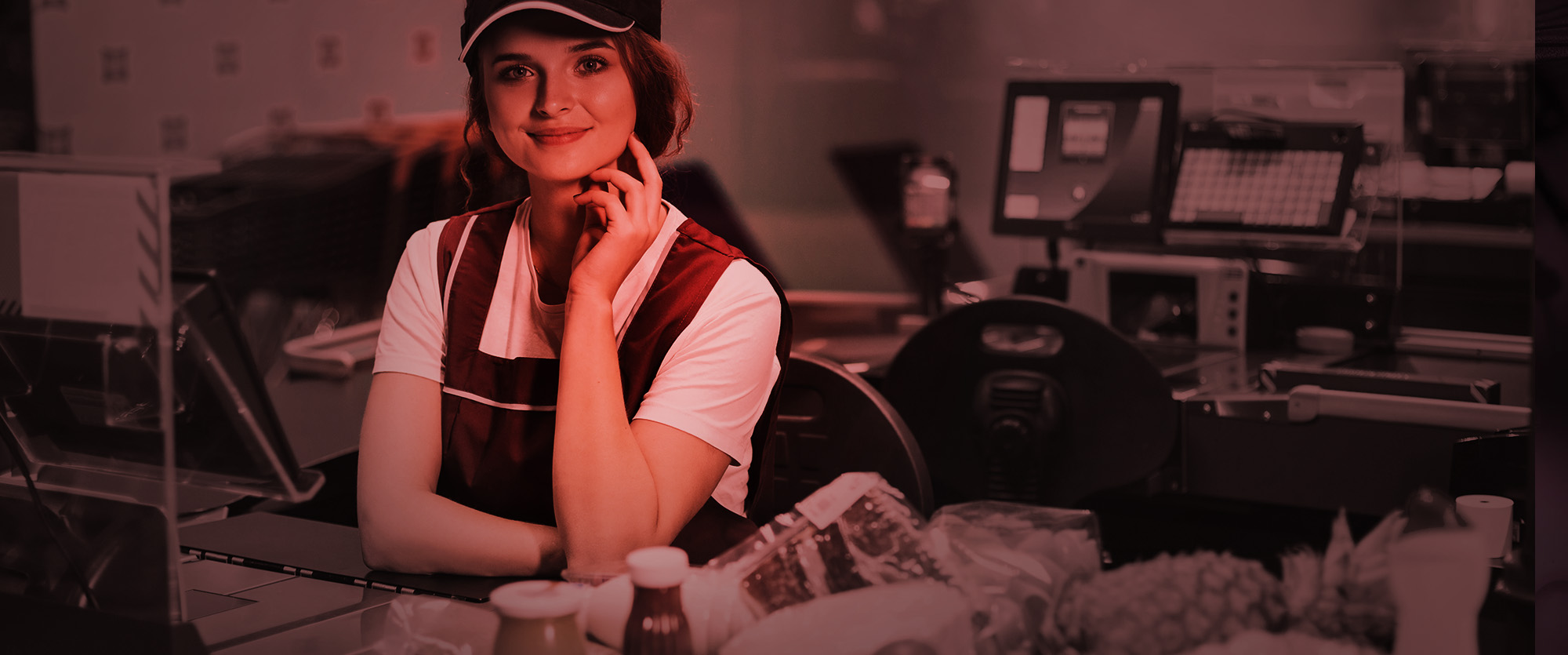 Smiling cashier in a maroon vest and cap seated at a convenience store checkout lane