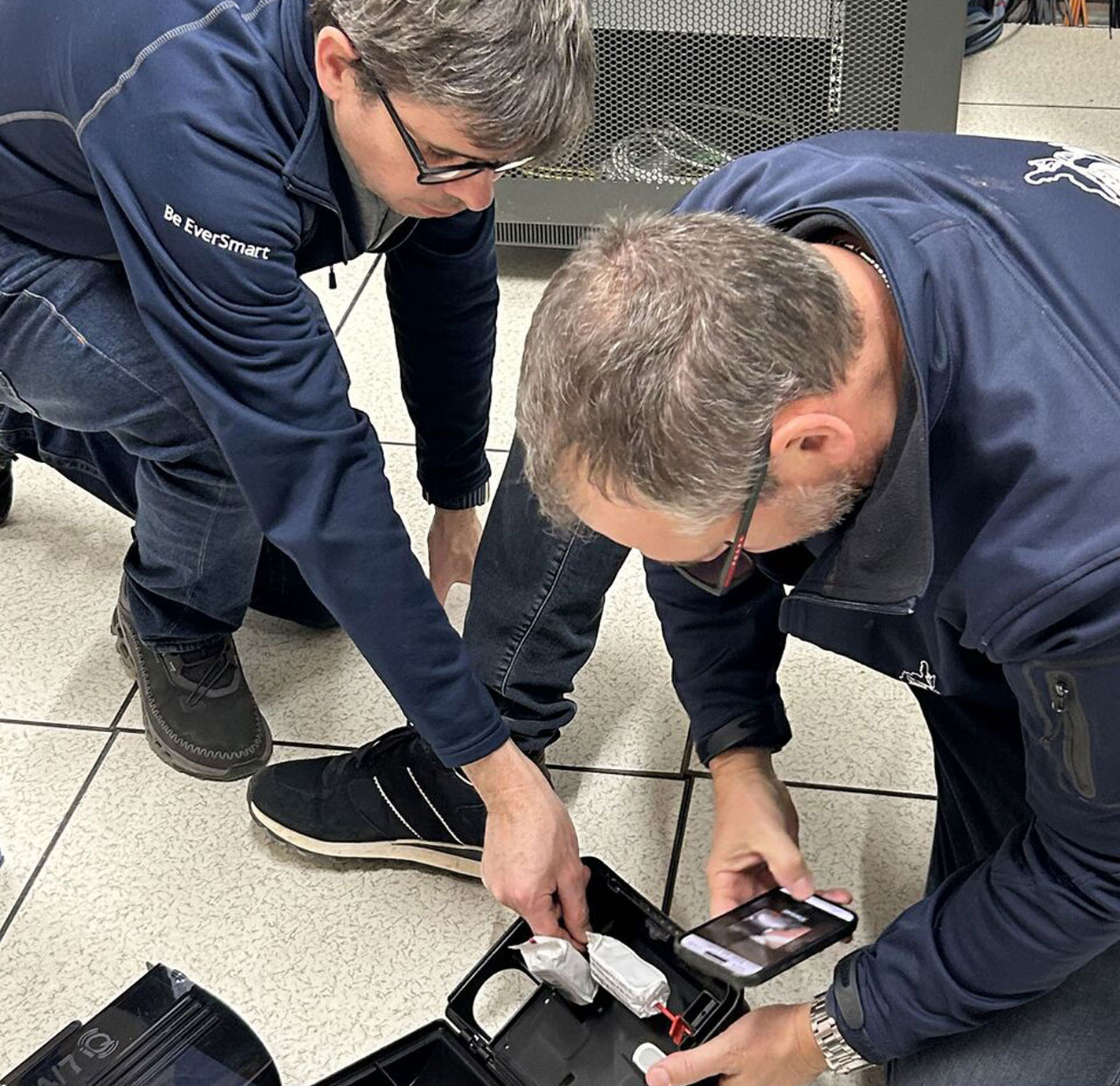 Two EverSmart technicians kneeling beside a server rack, fitting a monitored device from a carry case and photographing it with a smartphone